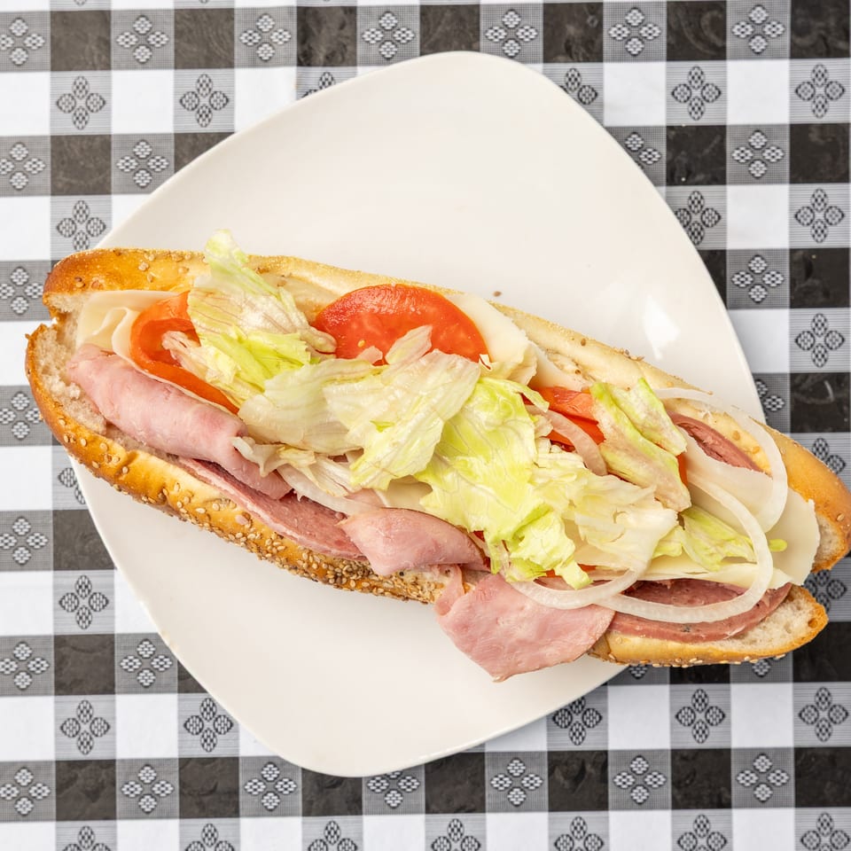 Sesame-seeded sandwich filled with ham, cheese, lettuce, tomato, and onion on a white plate atop a checkered tablecloth.