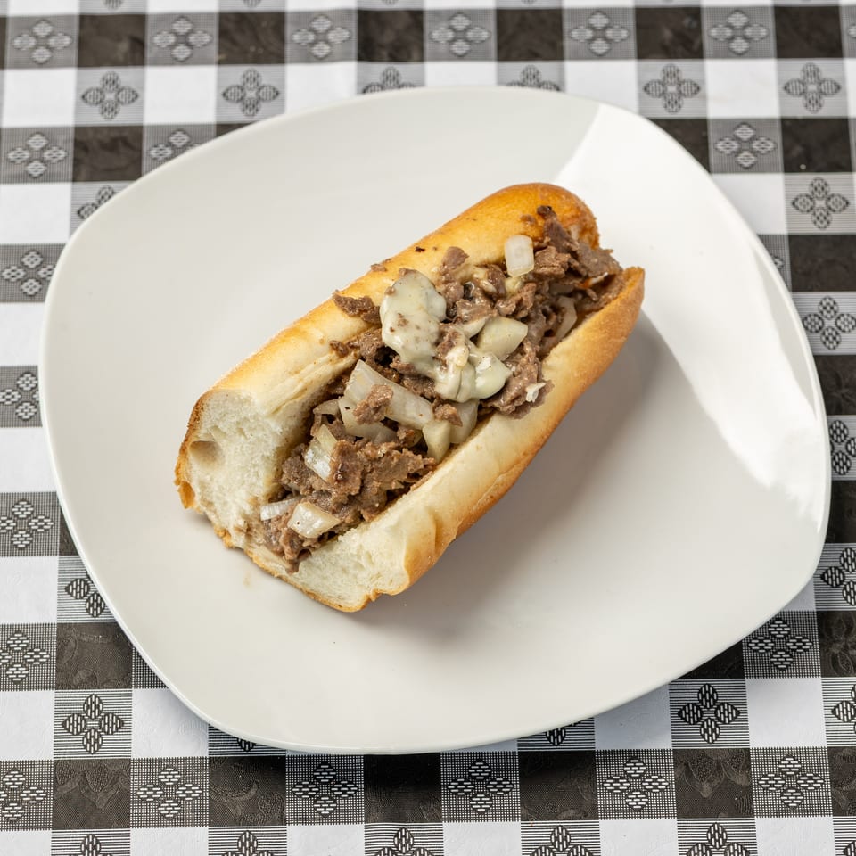 Philly cheesesteak sandwich with chopped beef, melted cheese, and onions on a white plate atop a checkered tablecloth.