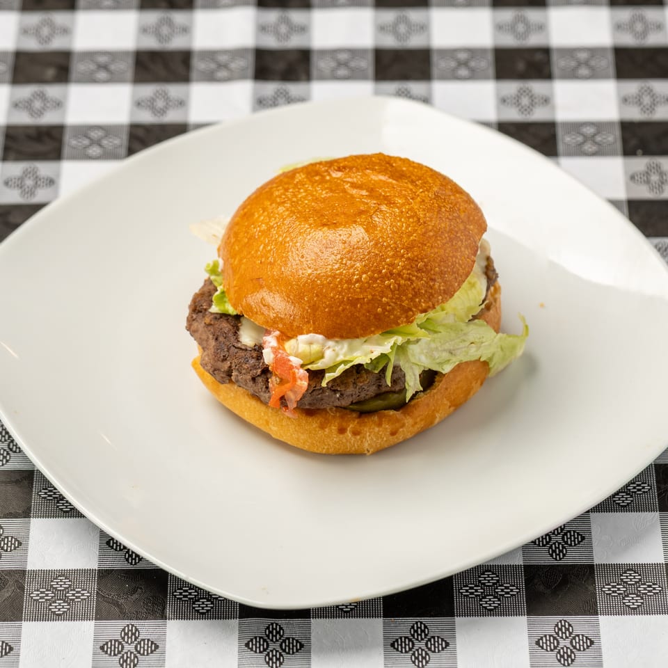 Juicy beef burger with lettuce, tomato, pickles, and creamy sauce on a white plate atop a black-and-white checkered tablecloth.
