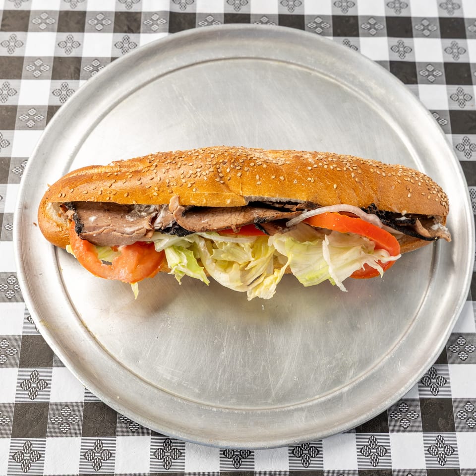 Sesame-seed sandwich filled with sliced roast meat, lettuce, tomato, onion, and sauce on a metal platter.